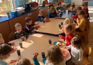 6 urodziny Nikoli. Dzieci siedzą przy trzech złączonych stołach. Jedzą tort. Obok talerzyków stoją kubeczki z sokiem.